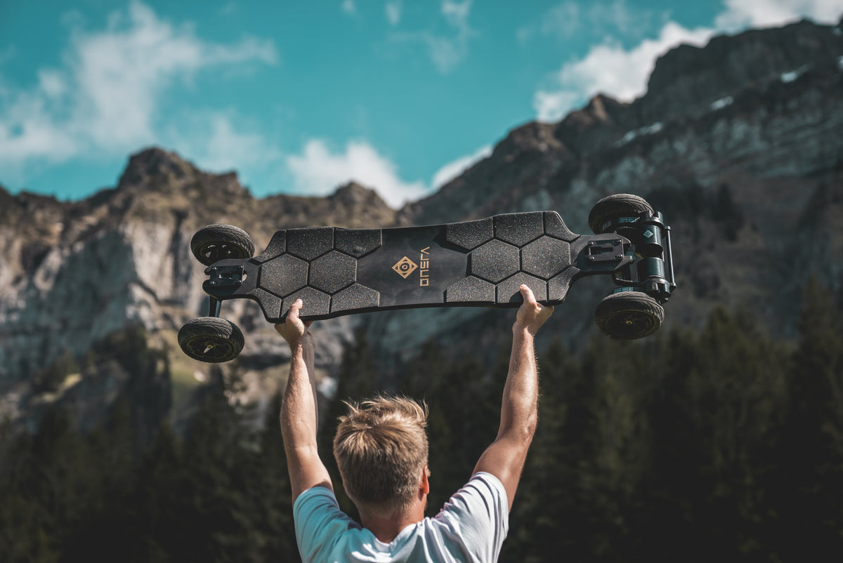 Person holding an ONSRA electric skateboard overhead in front of alpine mountains